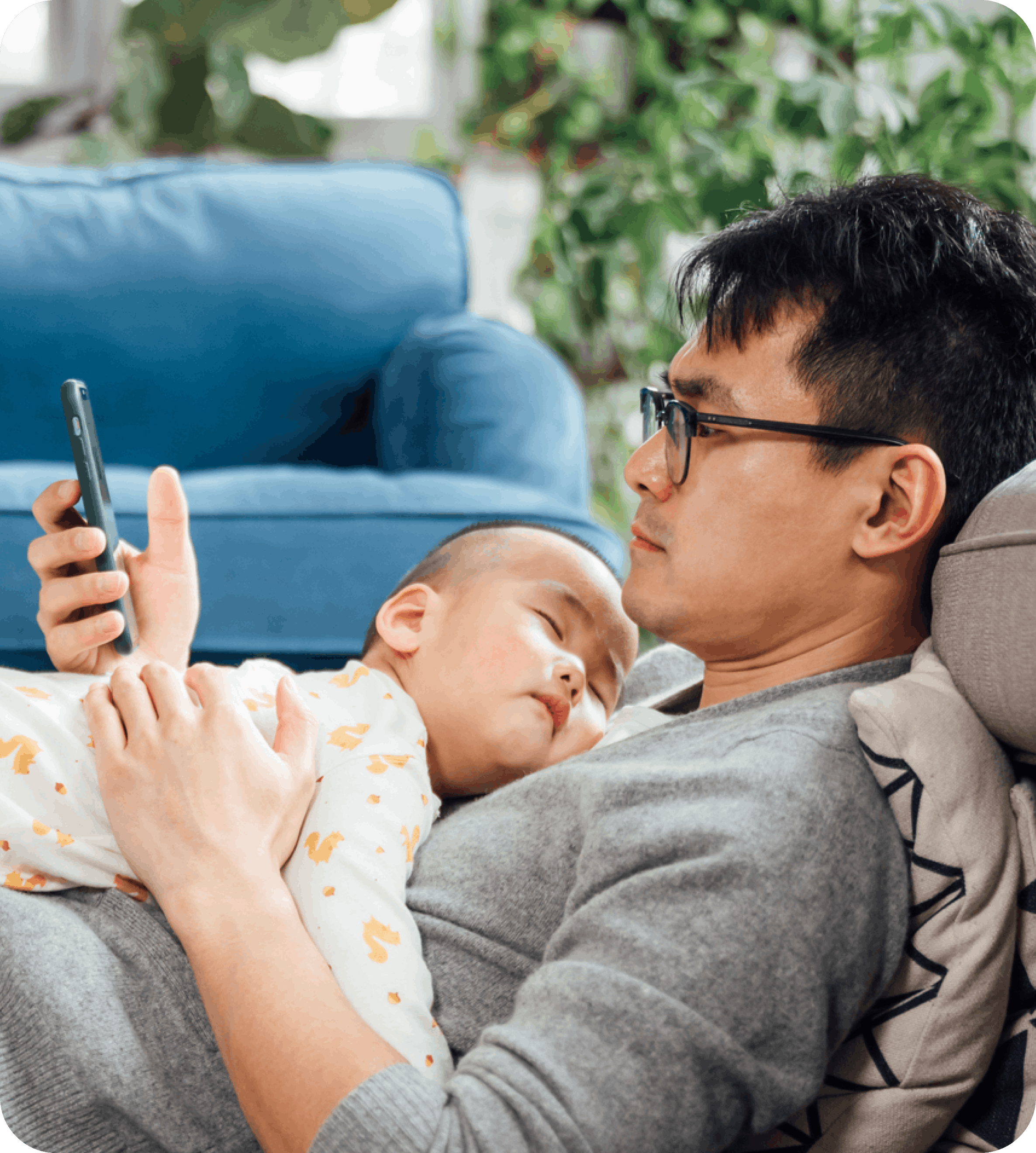 Image of a father holding his sleeping baby while looking at his smartphone at home.